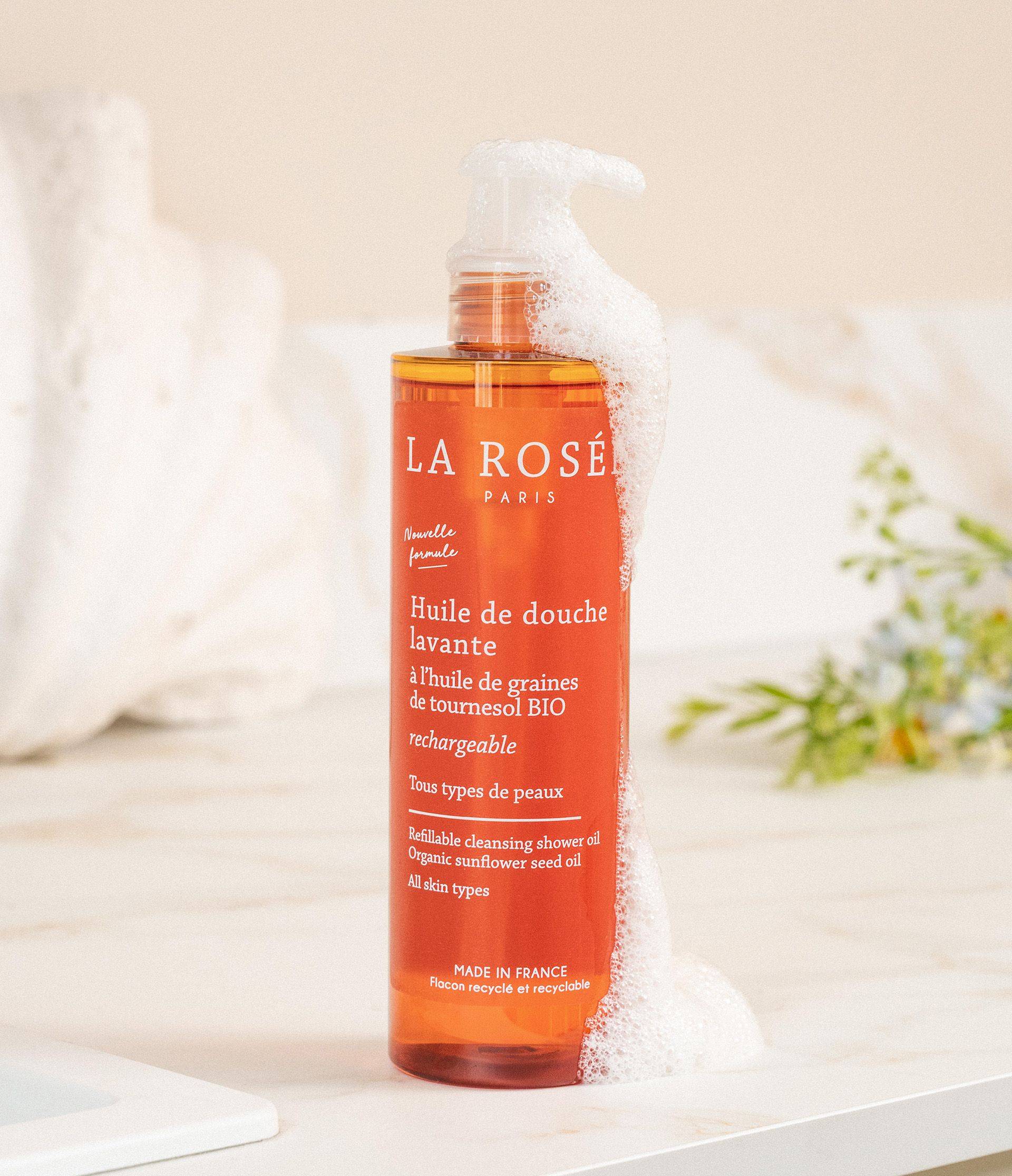 An amber bottle of La Rosé cleansing shower oil with white foam on a bathroom surface, surrounded by subtle plant decor.