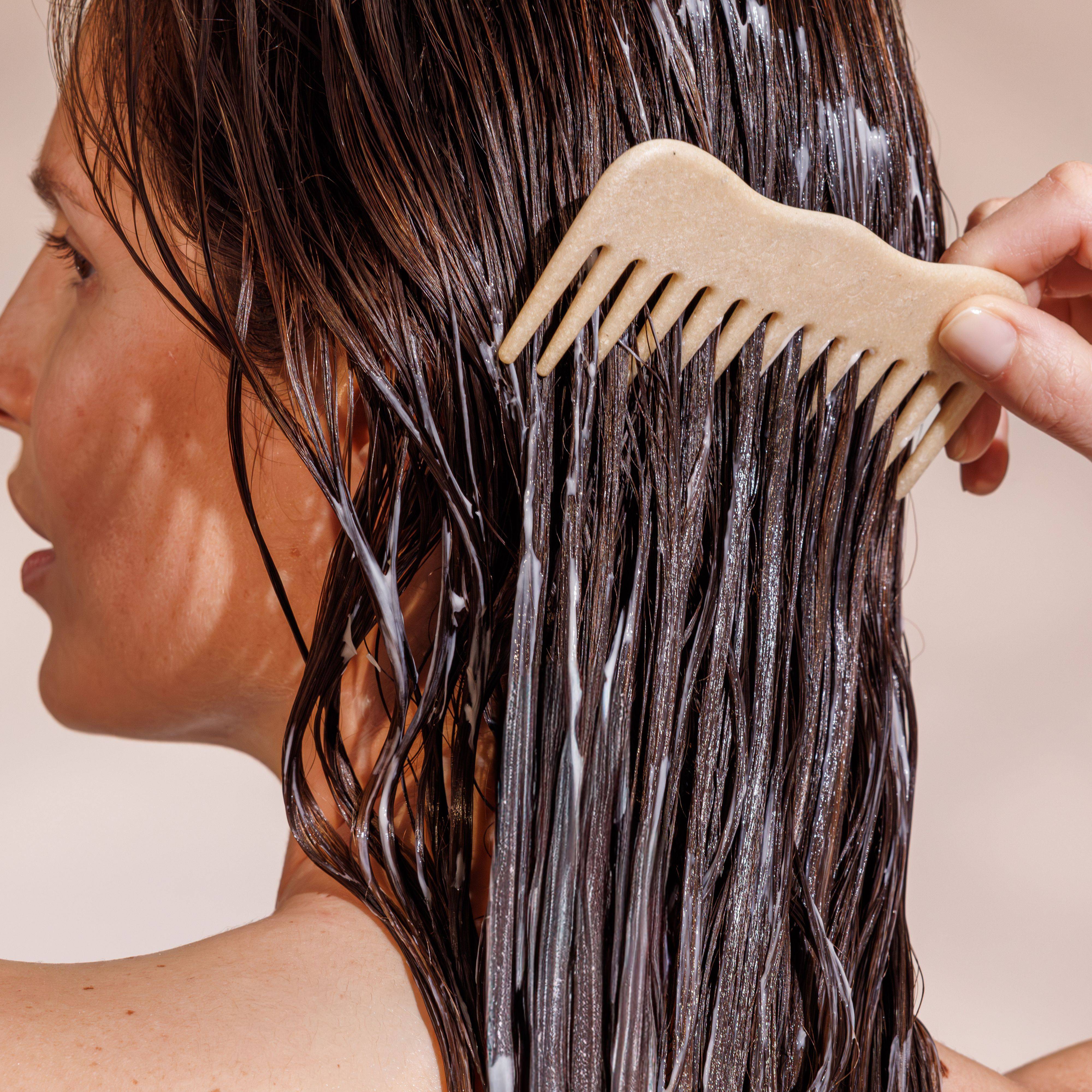 A person with wet hair is combing through it with a wide-toothed comb, applying conditioner.